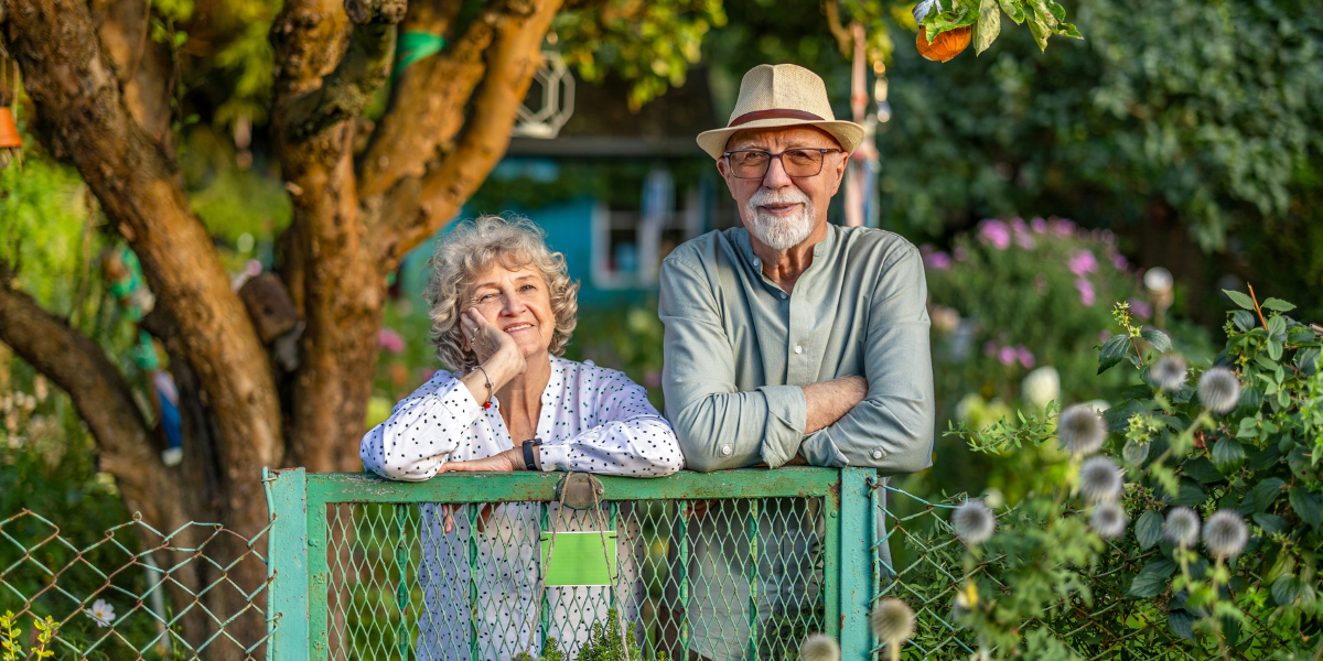 An older couple smiling warmly over a garden gate surrounded by lush greenery
